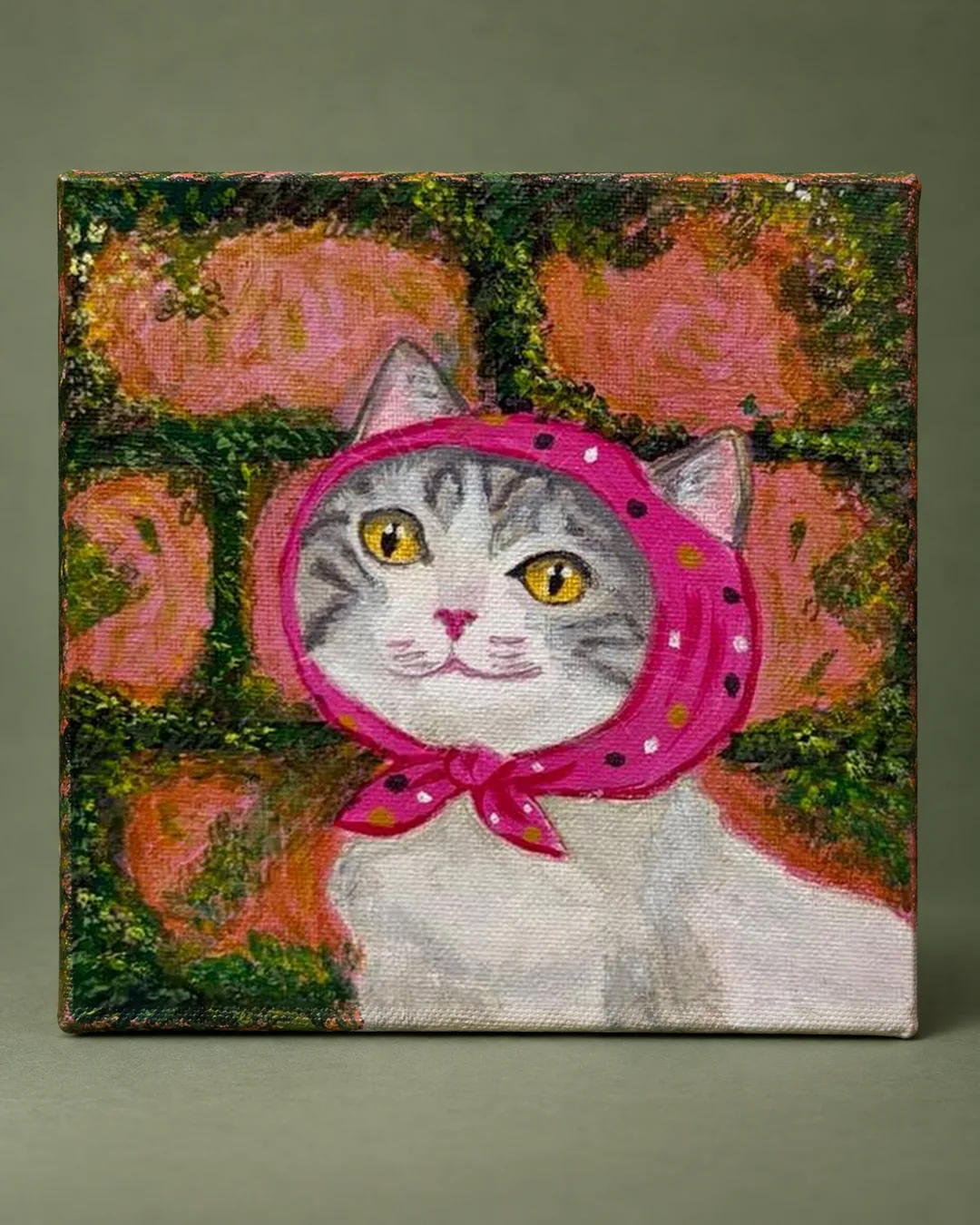 Sunday Best: a small painting of a grey tabby cat wearing a pink polka-dot headscarf in front of a mossy brick wall, photographed against a muted sage-green backdrop.