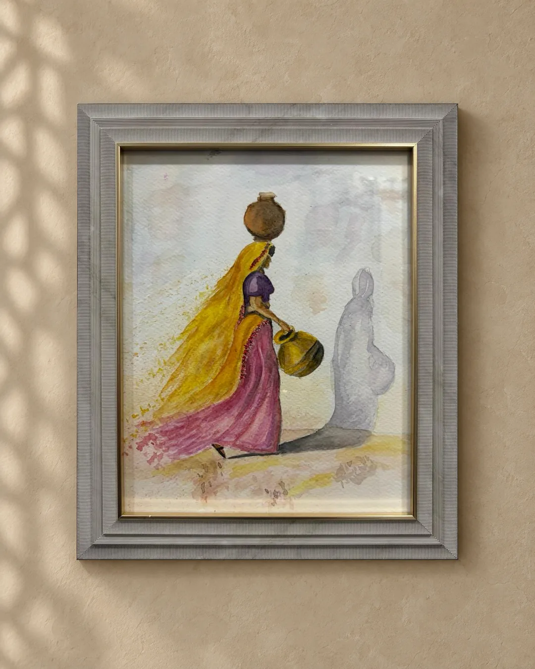 The Water Bearer: a framed watercolour of a woman in a saffron veil and rose skirt carrying clay water pots, hung on a warm sandstone wall with a soft lattice shadow.
